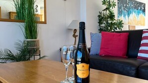 A wooden table with a bottle of Prosecco and two glasses in a living room with a black leather sofa, red and white striped pillows, and a large mirror on the wall.