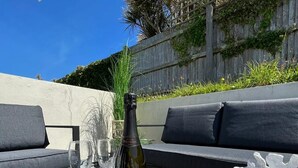 A bottle of Prosecco on a table with two glasses, a white coral sculpture, and a grey outdoor seating area with a wooden fence and greenery in the background.