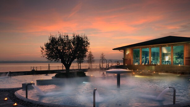A hot spring area with a large tree, a building with large windows, and a view of the water. - Modern apartment, Residence with swimming pool surrounded by greenery with FREE Wi-Fi (Sirmione)
