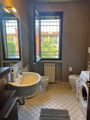 Een badkamer met een wastafel, toilet en wasmachine, een raam met uitzicht op gebouwen en een betegelde vloer.
