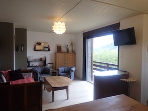 Ein Wohnzimmer mit einem Holztisch, zwei gemusterten Stühlen, einem Sofa, einer Holzkommode und einem an der Wand befestigten Fernseher.