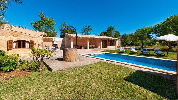 A poolside area with a stone barbecue, lounge chairs, and a building with a tiled roof. - Villa Els Olivers / Olivers Petits (Alcúdia)