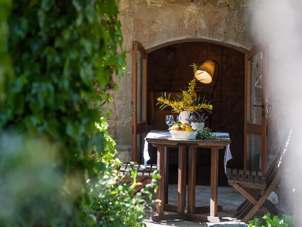 A rustic outdoor dining area with a wooden table set for two, a vase with yellow flowers, and a basket of fruit.