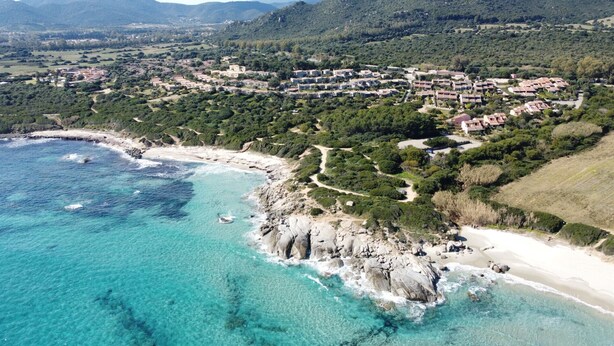 Vista aerea di una cittadina costiera con spiaggia limpida, costa rocciosa e un fitto gruppo di edifici.