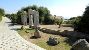 Un vialetto lastricato conduce a colonne in pietra antica, con grosse rocce e vegetazione circostante.