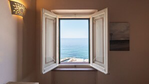 Een kamer met een raam dat uitkijkt op de zee, een wandlamp en een ingelijst schilderij.