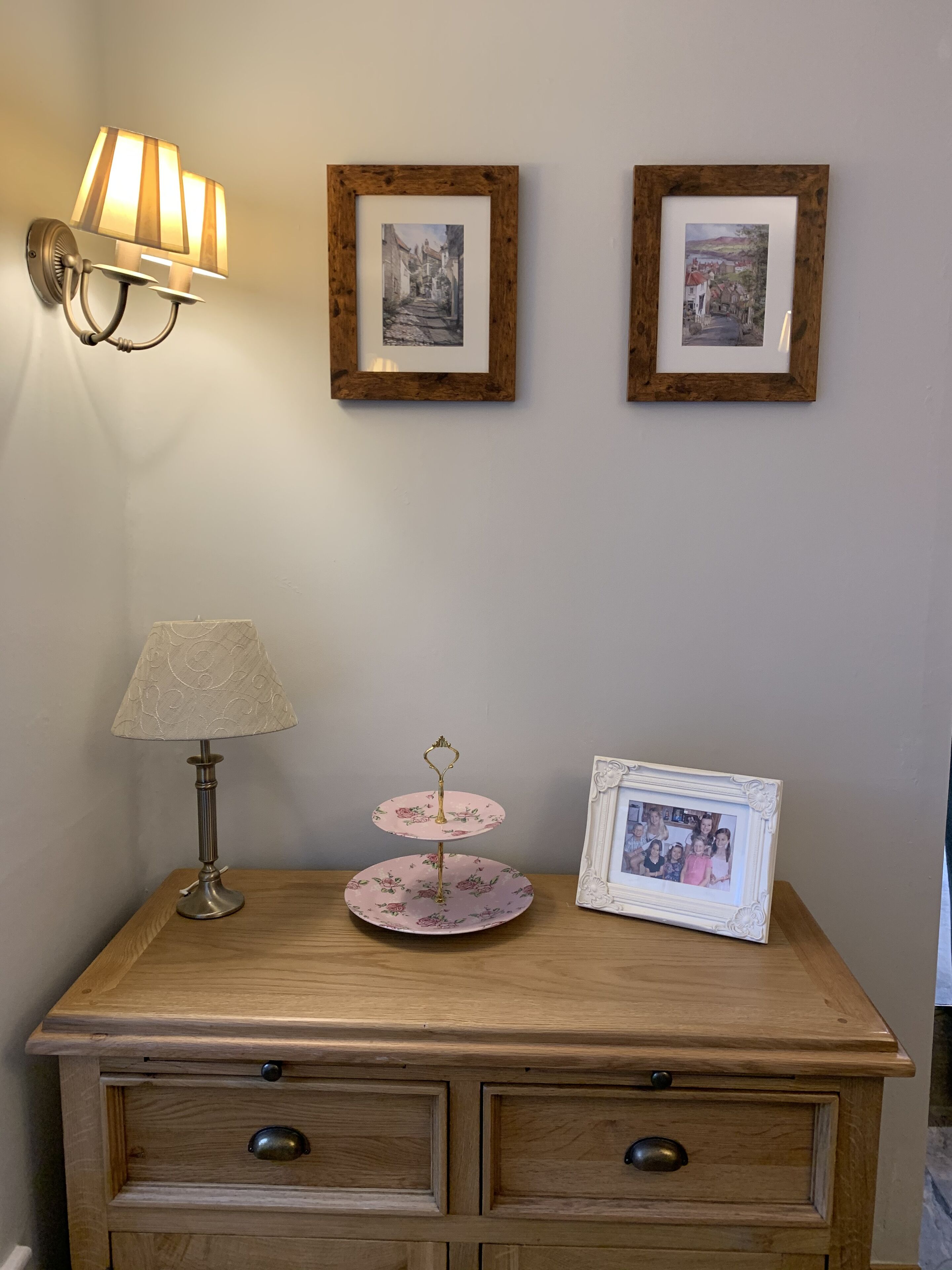 A wooden dresser with two drawers, a lamp, a pink floral cake stand, and two framed pictures on the wall.