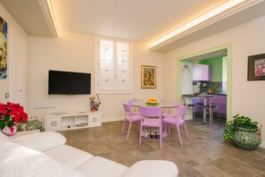 Een moderne woonkamer met een witte bank, een eettafel met glazen blad en paarse stoelen, een flatscreen televisie aan de muur en een zichtbare keuken door een open doorgang.
