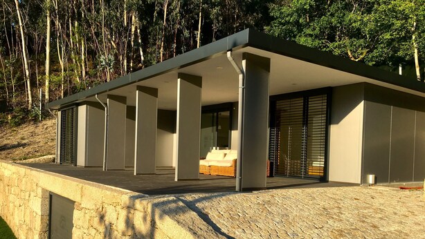 A modern house with a large balcony, a stone retaining wall, and a view of a forest.