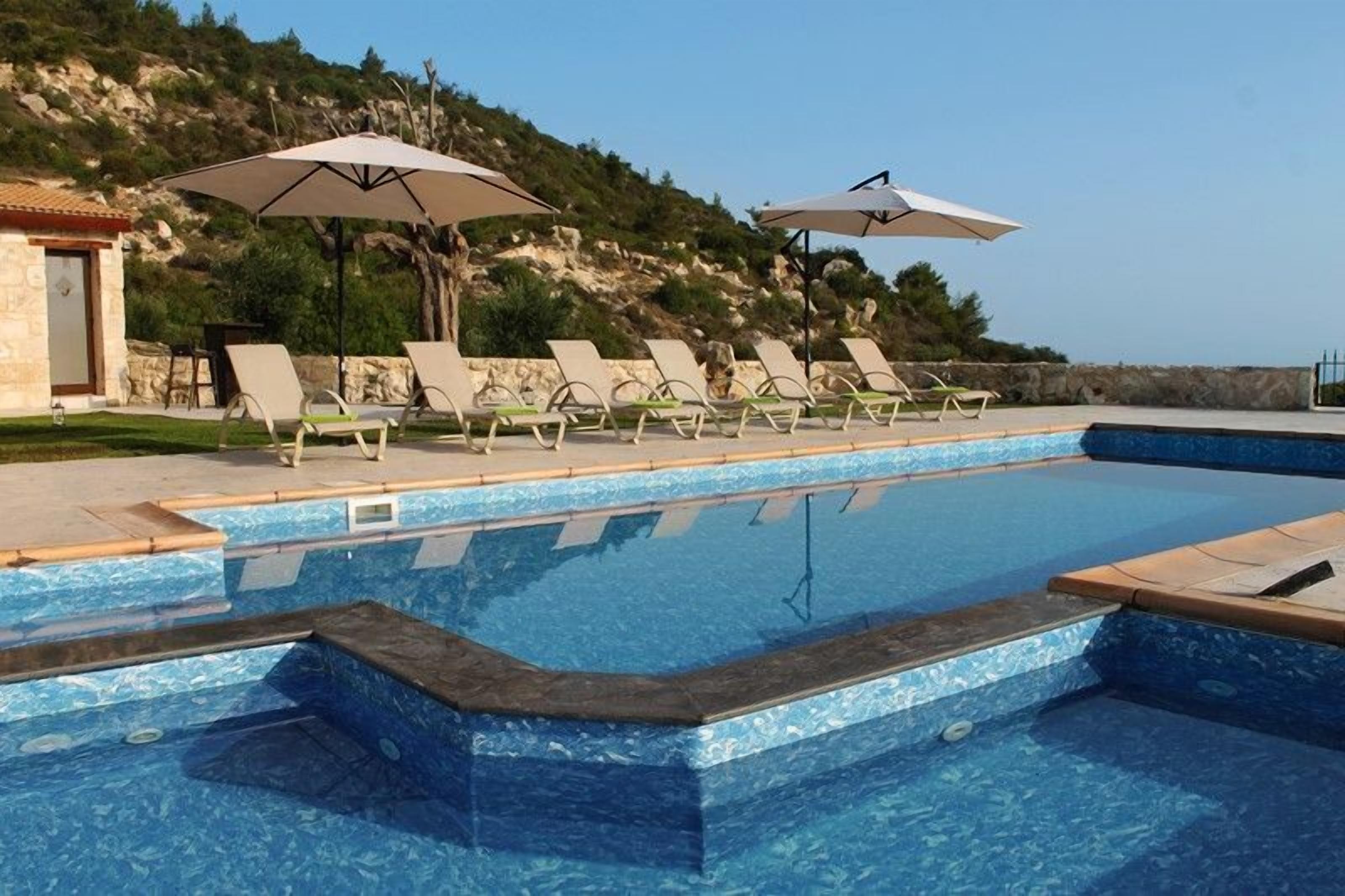 A swimming pool with lounge chairs and umbrellas, surrounded by a stone wall and a hillside.