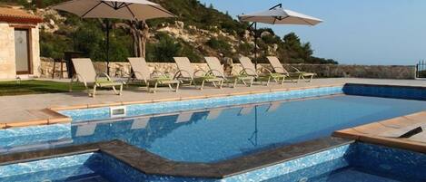A swimming pool with lounge chairs and umbrellas, surrounded by a stone wall and a hillside.