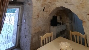 Um quarto com paredes de pedra, teto alto, luminária pendente, janela com grades metálicas e uma mesa com cadeiras.