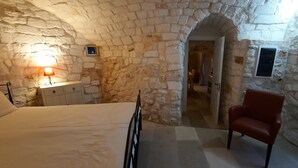 Quarto com paredes de pedra, uma cama, mesa de cabeceira com luminária, uma cadeira e uma porta aberta que dá para outro cômodo.