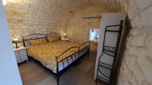 A bedroom with a stone wall, a bed with a patterned bedspread, a nightstand with a lamp, and a metal shelf unit.