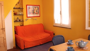Ein Zimmer mit gelber Wand, einem orangenen Sofa, einem Esstisch mit Teekanne und Tassen sowie einem Fenster mit Jalousien.