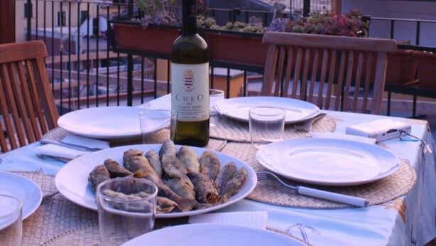 Ein Tisch ist für eine Mahlzeit gedeckt, mit Tellern, einer Weinflasche und einer Schüssel Essen auf einem Balkon mit Blick auf eine Stadtlandschaft.