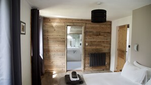 A modern hotel room with a wooden accent wall, a bed with white linens, a black ceiling light, and a view into the bathroom. - Popular, Stylish Chalet. Exceptional location with stunning Mont Blanc views. (Chamonix-Mont-Blanc)