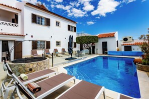 Zona junto a la piscina con sillones de descanso, una fogata de piedra y un cielo azul despejado.