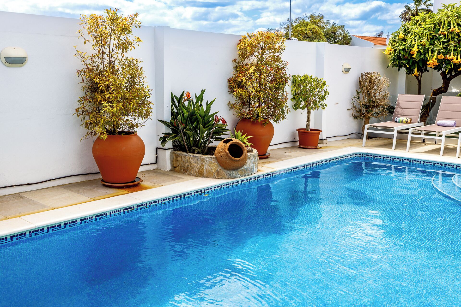A swimming pool with potted plants, a white wall, and a lounge area with chairs.