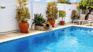 A swimming pool with potted plants, a white wall, and a lounge area with chairs.