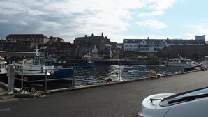 A harbor with boats docked, a car on the road, and buildings in the background. - Detached Cottage In Heart Of This Beautiful Harbour Village (Seahouses)