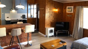 A modern living room with a wooden wall, a black sofa, a wooden coffee table, and a kitchen area with a white countertop and hanging lights. - Detached Cottage In Heart Of This Beautiful Harbour Village (Seahouses)