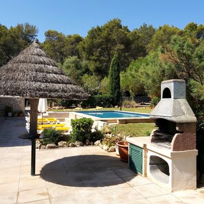A pool area with a stone barbecue and a thatched-roof structure. - House / Villa - Pollensa (Pollenca)