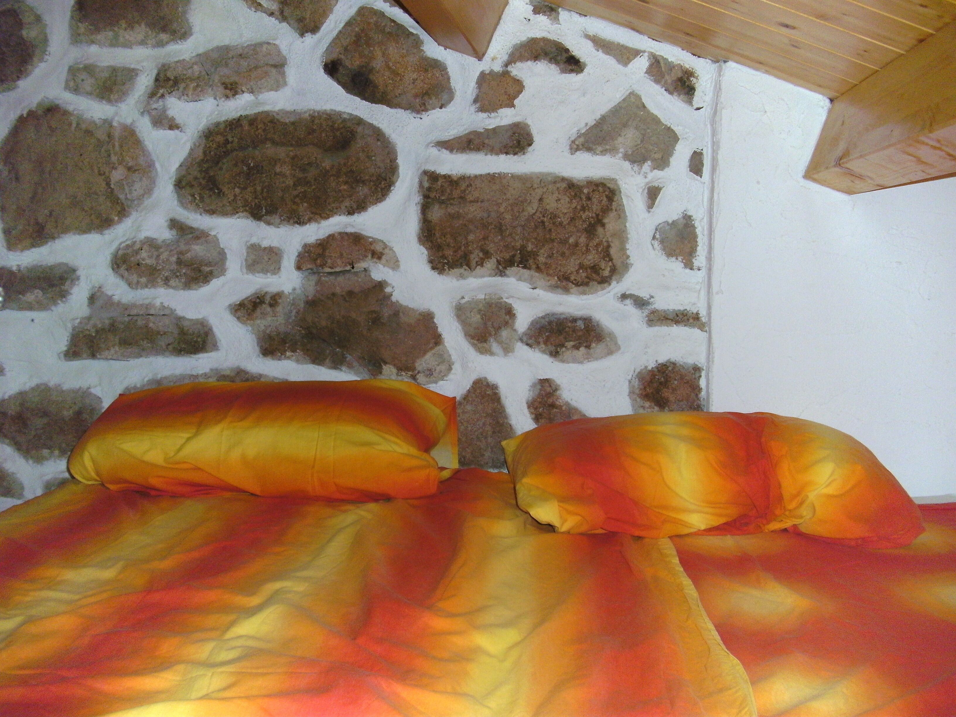 Ein Bett mit bunter Decke und Kissen, eine Steinwand und eine Holzbalkendecke.