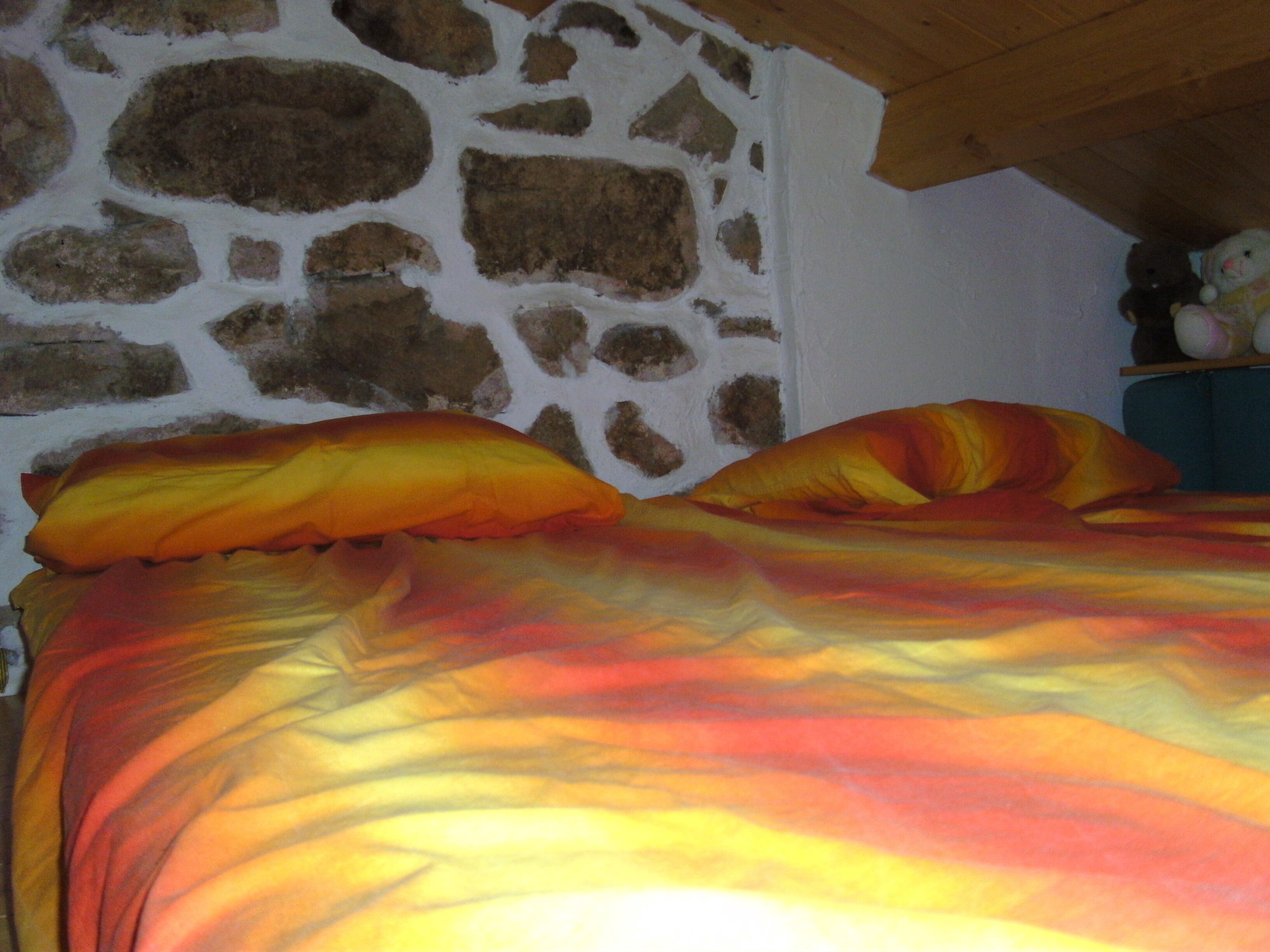 Ein Bett mit bunter Decke und Kissen, eine Steinsäule und ein Regal mit Stofftieren.