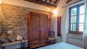 Een slaapkamer met een stenen muur, een houten garderobe, een bed met quilt, een stoel en een raam met luiken.