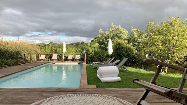 Una piscina con sillas de descanso y sombrillas, rodeada de vegetación y una terraza de madera.