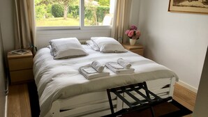 Una cama bien hecha con sábanas y almohadas blancas, una mesita de noche con una lámpara, una ventana con cortinas y un pequeño jarrón con flores.