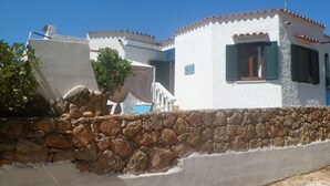 A white building with a stone wall, a staircase, and a window with green shutters. - Casa Niperu in the countryside a few meters from the beaches of Cala Ginepro (Cala Liberotto)