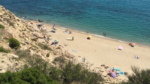 Una playa con aguas azules cristalinas, orilla arenosa y terreno rocoso.