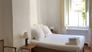Un dormitorio con cama, una silla, una mesita de noche, una lámpara y una ventana con cortinas.