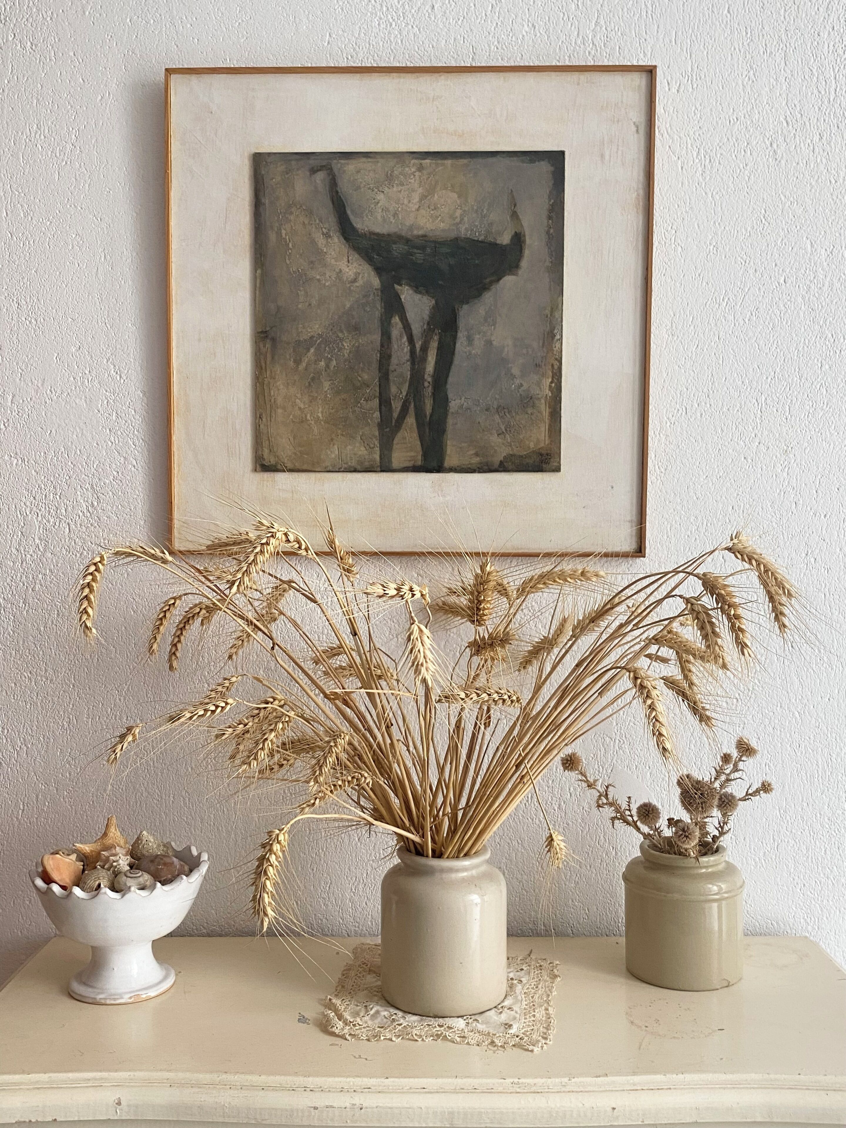 A framed abstract painting on a textured wall, a vase with dried wheat stalks, and a small bowl with seashells on a wooden shelf.