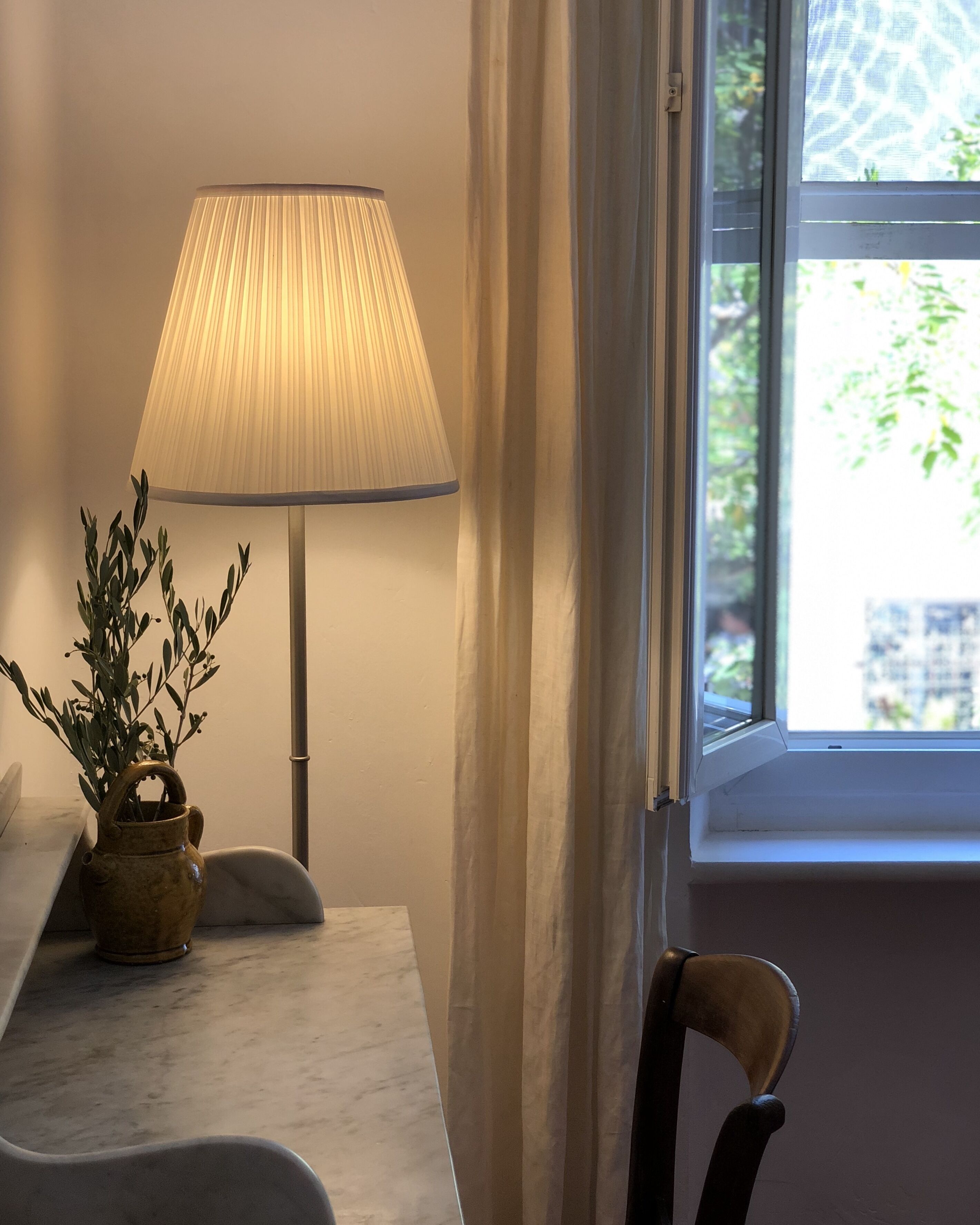 Une pièce avec une table, une lampe, un vase avec une plante et une fenêtre avec des rideaux.