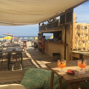 An outdoor seating area with tables and chairs, a bar counter, and a view of the beach.