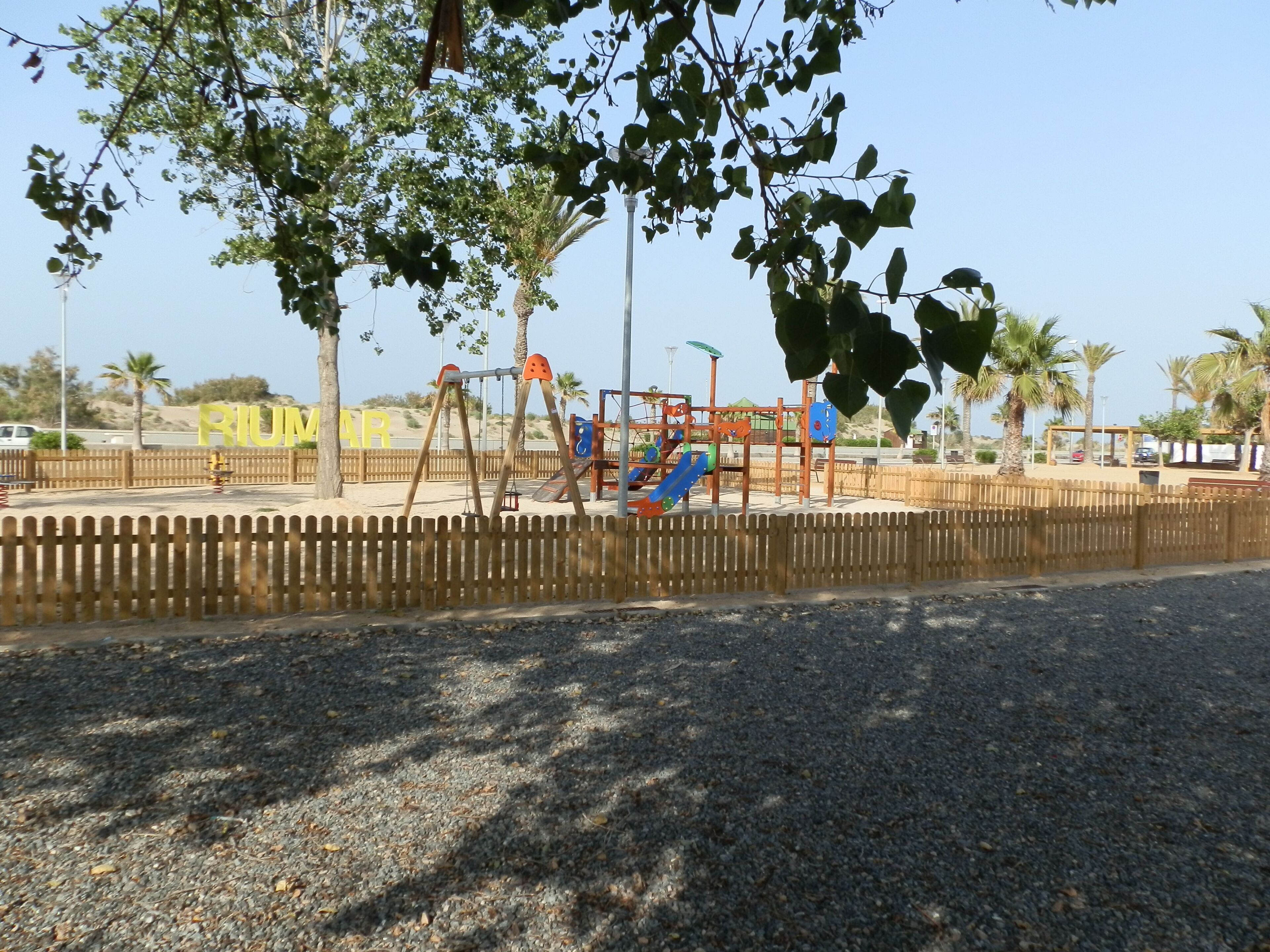 A playground with swings and slides, surrounded by a wooden fence and palm trees.