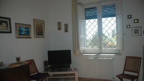 A living room with a TV, a dining table, and chairs. There are framed pictures on the wall.