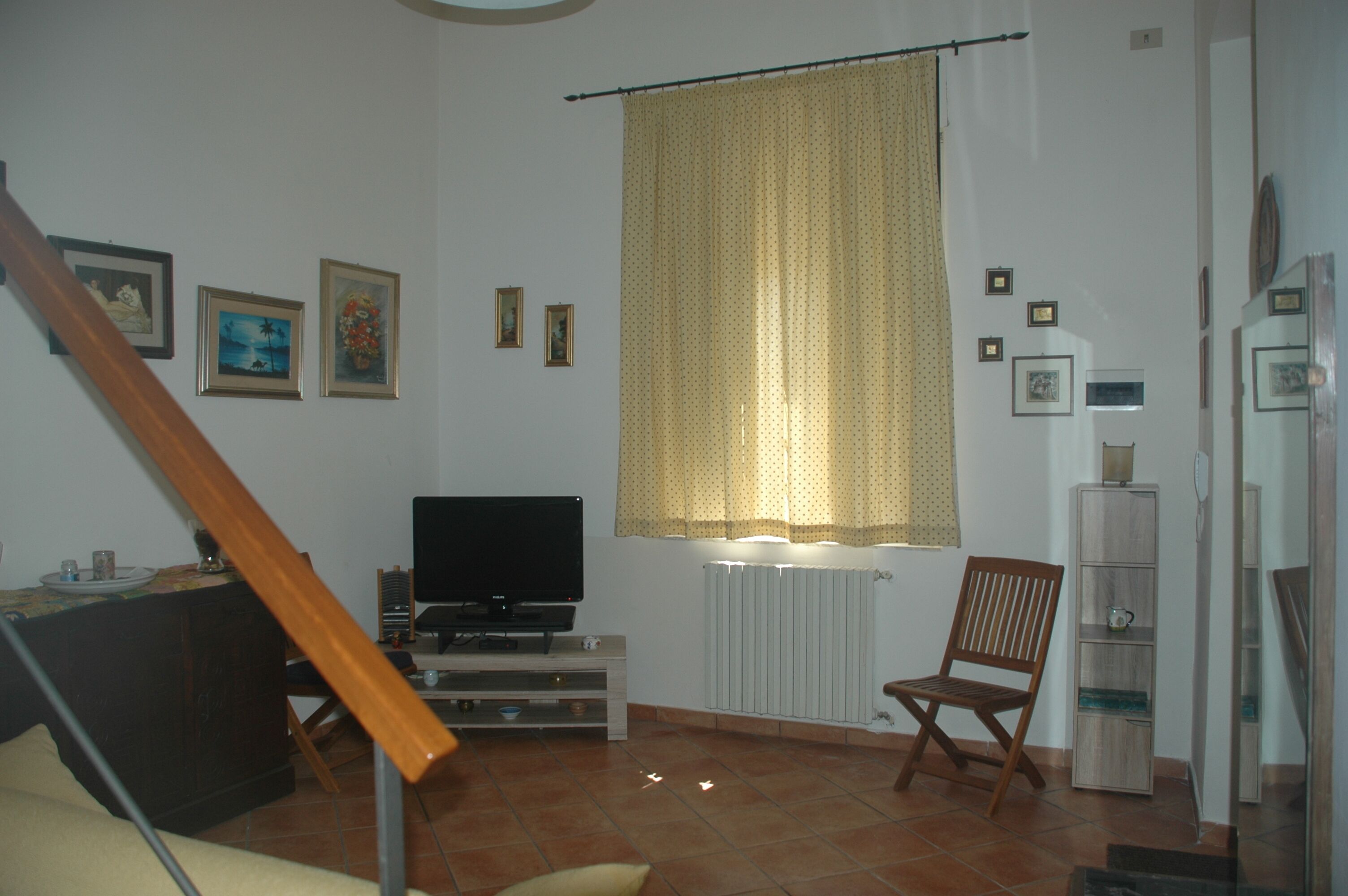 Un salon avec un téléviseur, une chaise en bois, un radiateur et une fenêtre avec des rideaux.