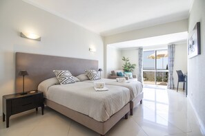A bedroom with two beds, a nightstand, a lamp, a sofa, and a balcony with curtains.