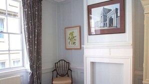 Ein Zimmer mit Kamin, einem gerahmten Bild, einem Sessel und einem Fenster mit Vorhängen.
