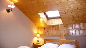 Een slaapkamer met een schuin houten plafond, dakraam, bed met witte lakens, nachtkastje met lamp en een wandlamp.