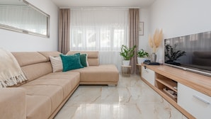 A modern living room with a beige sofa, a flat-screen TV, a marble floor, and a large window with sheer curtains.