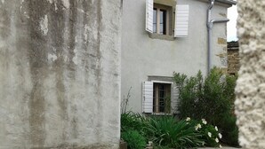 Un bâtiment dont l’extérieur est marqué par les intempéries, avec une petite fenêtre ornée de volets et un jardin planté de fleurs et d’autres végétaux.