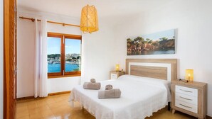 Une chambre à coucher avec un grand lit, une tête de lit en bois, une table de chevet avec une lampe, et une vue sur une ville côtière par la fenêtre.