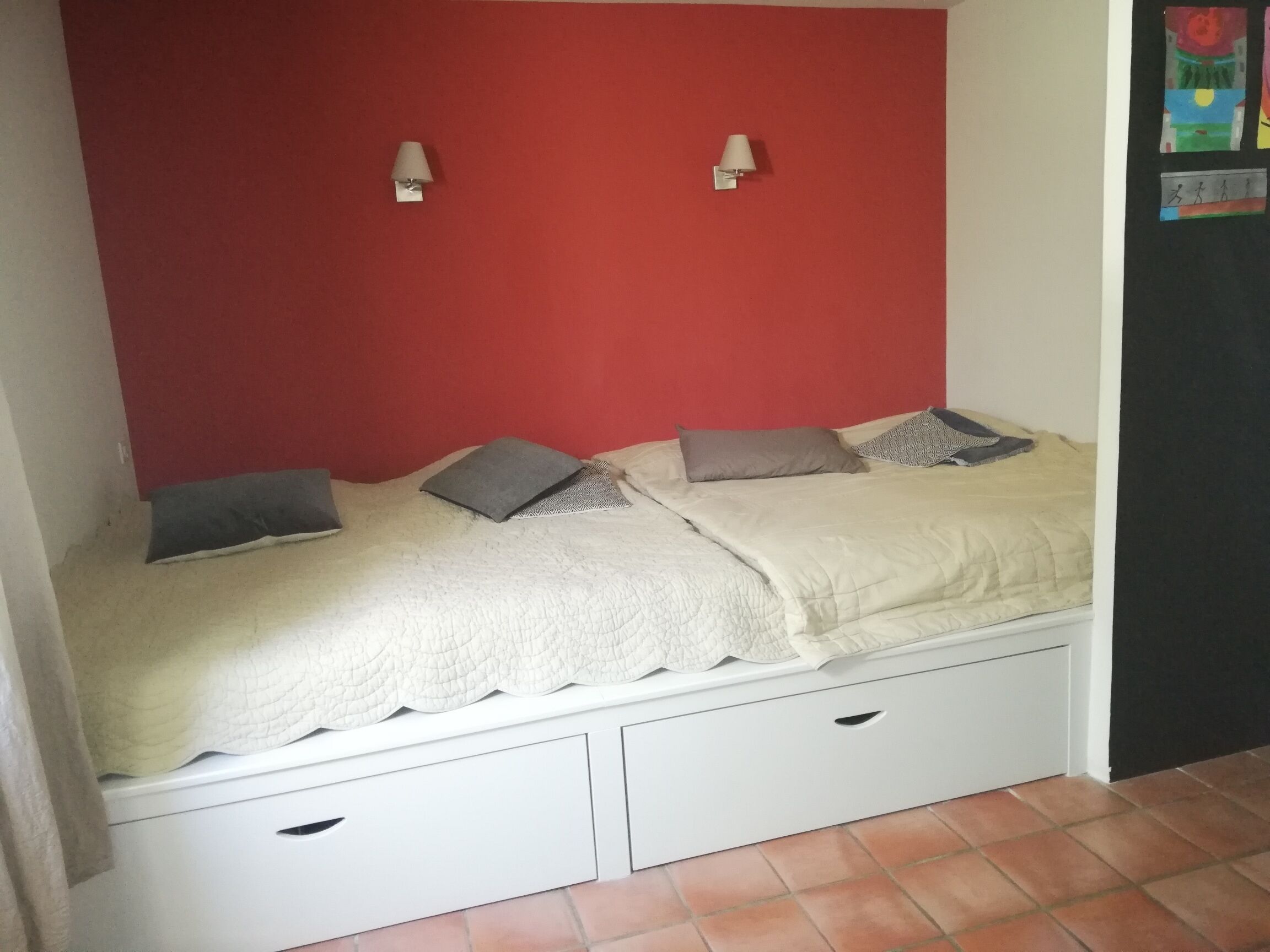 A small bedroom with a red wall, two bedside tables, a bed with a quilted cover, and two wall-mounted lamps.