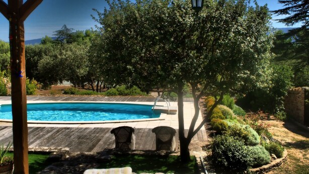 A wooden deck with a swimming pool, wicker chairs, and a pergola with a hanging light fixture. - A cozy home surrounded by nature, with a private pool and tennis court (Cabrières-D'avignon)