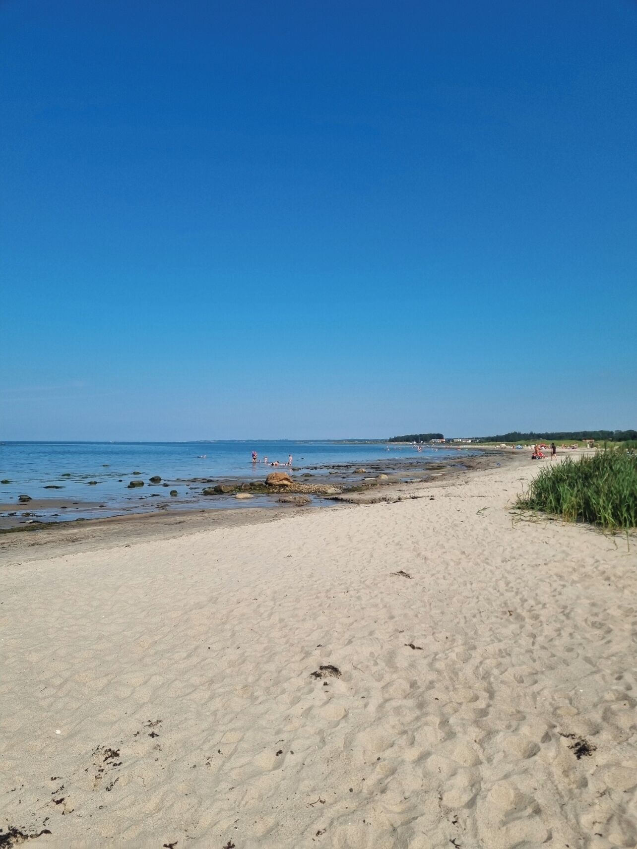 A sandy beach with clear blue water, some rocks, and a few people in the distance.